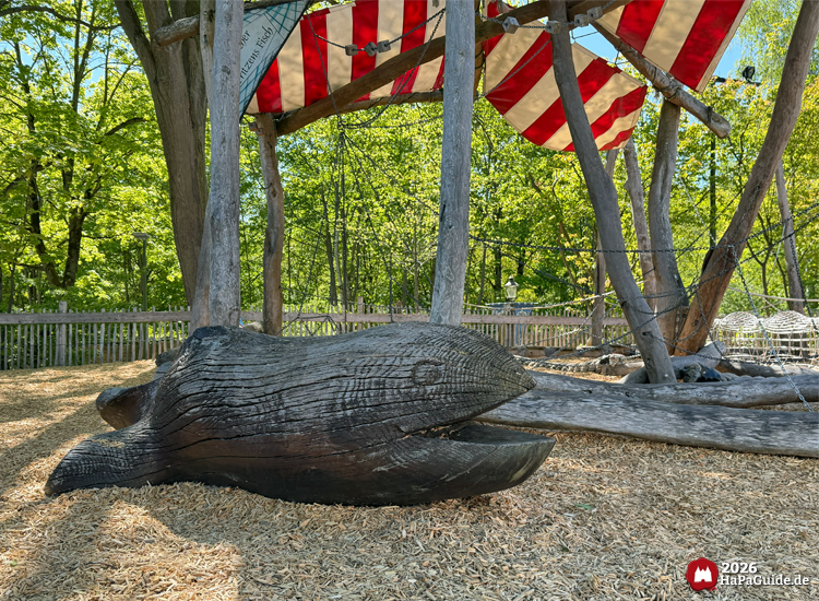 Große Holzskulptur eines Wals im Spielbereich Niedrigseilgarten im Hansa-Park, unter roten und weißen Segeltüchern, umgeben von Holzspänen und Bäumen