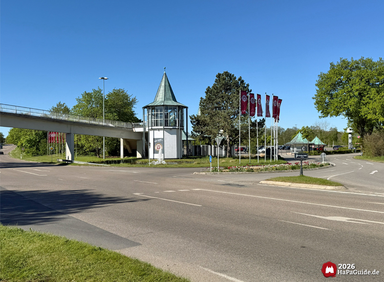 Einfahrt vom Hansa-Park mit oktogonalem Turm, Fußgängerbrücke über der Zufahrtsstraße und wehenden Hansa-Park Fahnen an einem sonnigen Tag