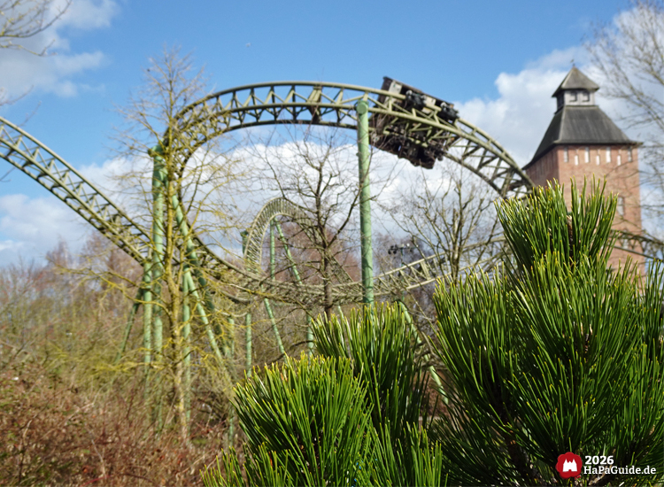 Achterbahn Flucht von Novgorod im Hansa-Park mit grünem Schienensystem und Zug im Buchtknoten, umgeben von kahlen Bäumen und dem Backsteinturm Saviour-Tower unter blauem Himmel
