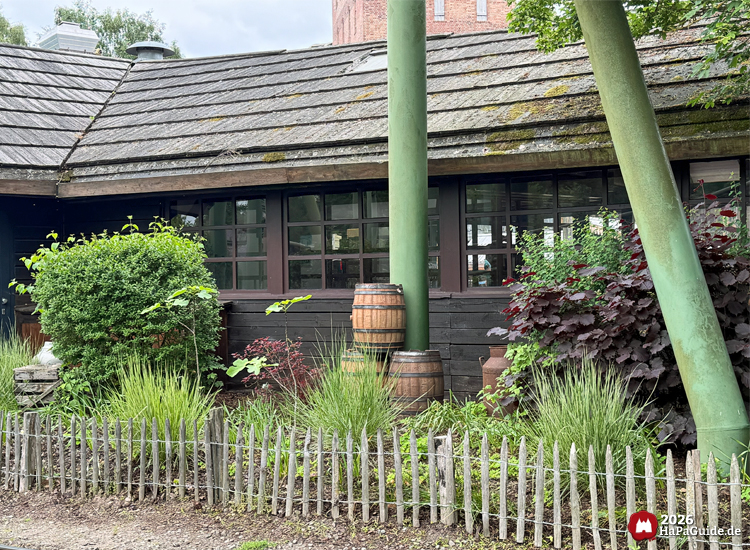 Rückseitiger Kiosk im Peterhof im Hansa-Park mit dunkler Holzfassade, begrüntem Garten und Holzfässern, im Hintergrund der markante Backsteinturm der Achterbahn Flucht von Novgorod