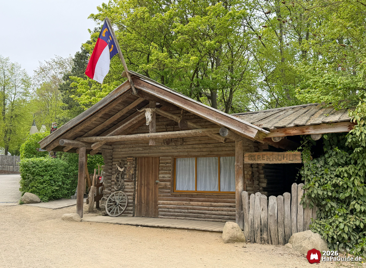 Rustikale Blockhütte Kiosk Tipi-Lager im Hansa-Park mit Holzfassade, Flagge von North Carolina und Wagenrad-Dekoration vor grünen Bäumen
