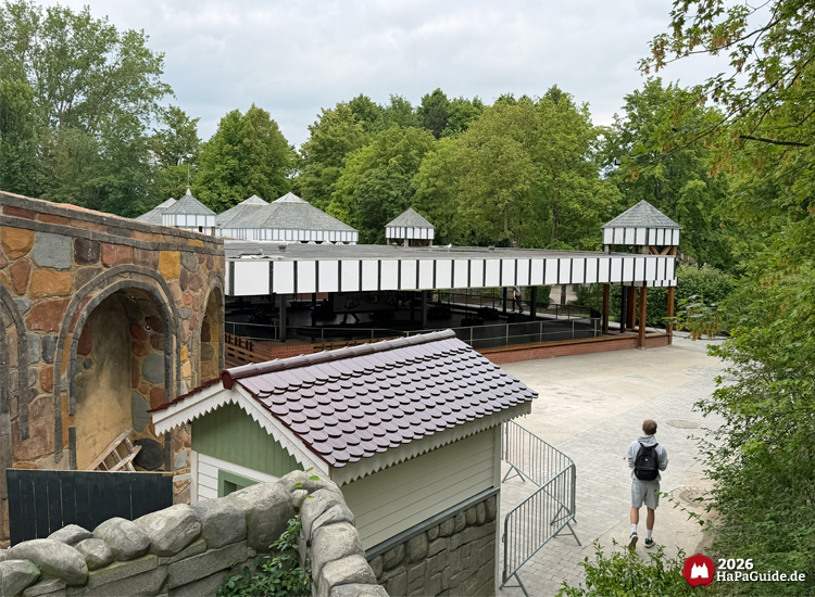 Baustellenbereich am Cornwall Coaster im Hansa-Park mit weiß-schwarzem Gebäude von 1903 Somerset Traffic, Steinmauer und kleinem grünen Häuschen, umgeben von Bäumen und Wegen