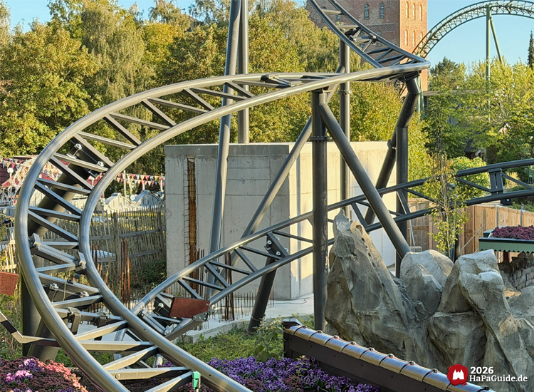 Stahlkonstruktion der Achterbahn Cornwall Coaster im Hansa-Park mit geschwungener Schiene und Betonwand, umgeben von Felsen, Blumen und grünen Bäumen im Hintergrund