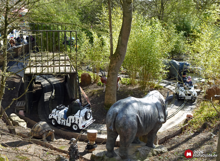 Zebra-Jeep der Safari-Tour im Hansa-Park fährt durch eine Dschungellandschaft mit großer Nashornfigur, Bambus und Bäumen, im Hintergrund ein Holzsteg und weitere Jeeps