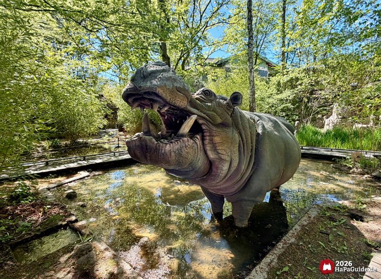 Große Nilpferdfigur mit geöffnetem Maul im Wasserbecken der Safari-Jeeps im Hansa-Park, umgeben von Bäumen und dichtem Grün unter blauem Himmel