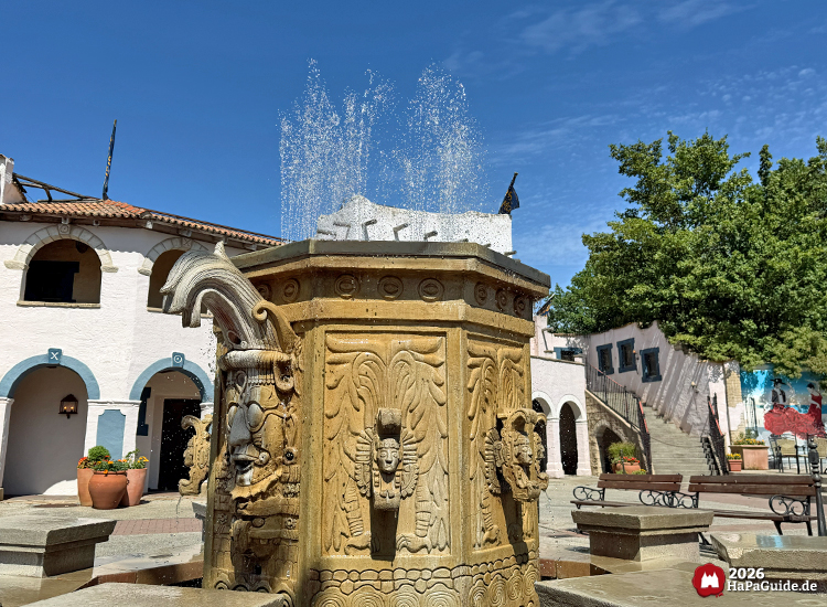 Steinbrunnen mit kunstvollen Reliefs und Wassersprühern im Themenbereich Fiesta del Mar des Hansa-Parks, umgeben von weißen Gebäuden mit blauen Bögen und Terrakottatöpfen