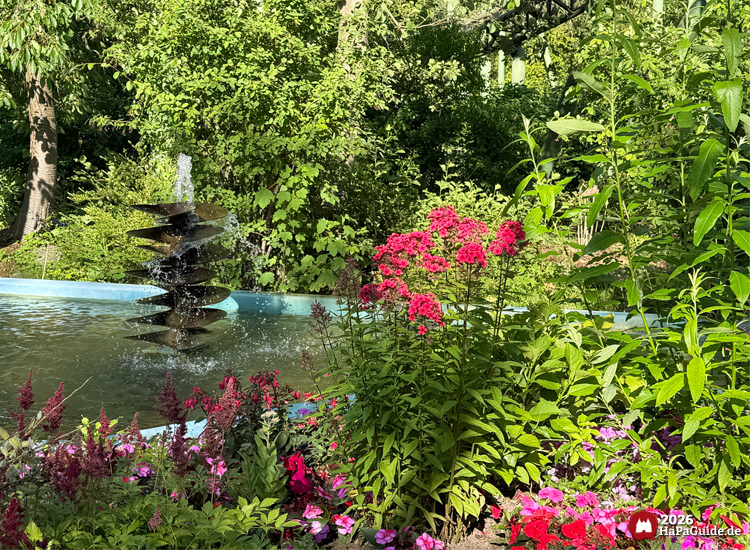Schiffsschraube als Brunnen im Hansa-Park mit Wasserstrahl, umgeben von pinken und roten Blumen sowie dichtem Grün, sonniger Sommertag