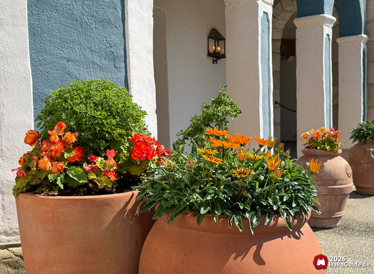 Große Terrakottatöpfe mit roten Begonien und orangefarbenen Blüten vor weiß-blauer Fassade im Themenbereich Fiesta del Mar des Hansa-Parks, sonniger Tag mit klarer Beleuchtung