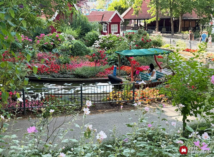 Blumenmeerbootsfahrt des Hansa-Parks mit grünem Dach über dem Boot, umgeben von roten, pinken und weißen Blumenbeeten sowie kleinen roten Häusern mit Ziegeldächern