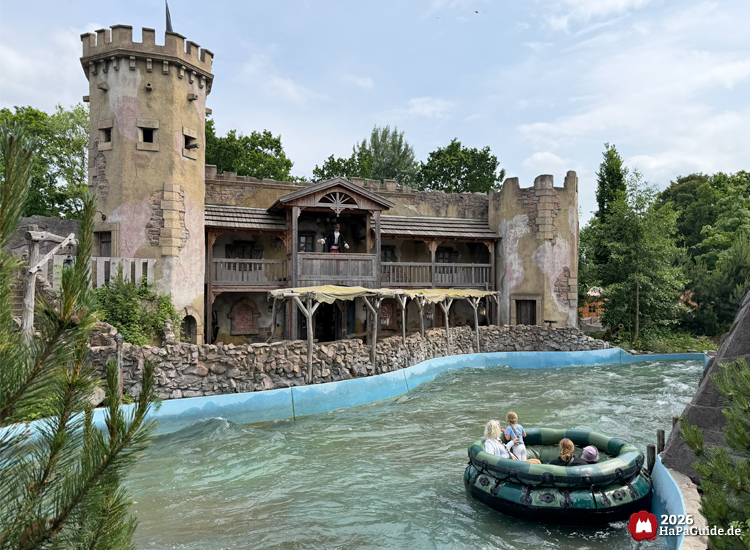 Burgkulisse der Wasserattraktion Störtebeker’s Kaperfahrt im Hansa-Park mit Turm, Holzveranda und Piratenfigur Störtebekers, Rundboot mit Fahrgästen auf der blauen Wasserbahn im Vordergrund
