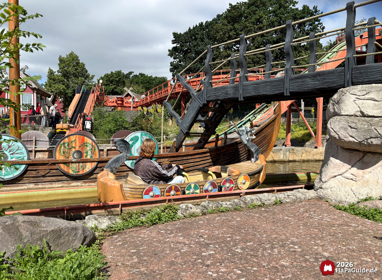Sturmfahrt der Drachenboote im Hansa-Park mit Wikingerschiff Viking Odin, verzierten Rundschilden und geschnitzten Drachenköpfen, unter einer schwarzen Holzbrücke mit Seilgeländer, umgeben von Felsen und grüner Landschaft