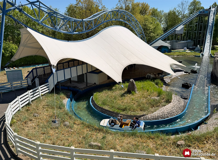 Wasserbahn Super Splash im Hansa-Park mit Boot auf Wasserkanal unter weißem Zeltdach, dahinter Lifthill und Wasserabfahrt, umgeben von grüner Landschaft und blauem Fahrkanal