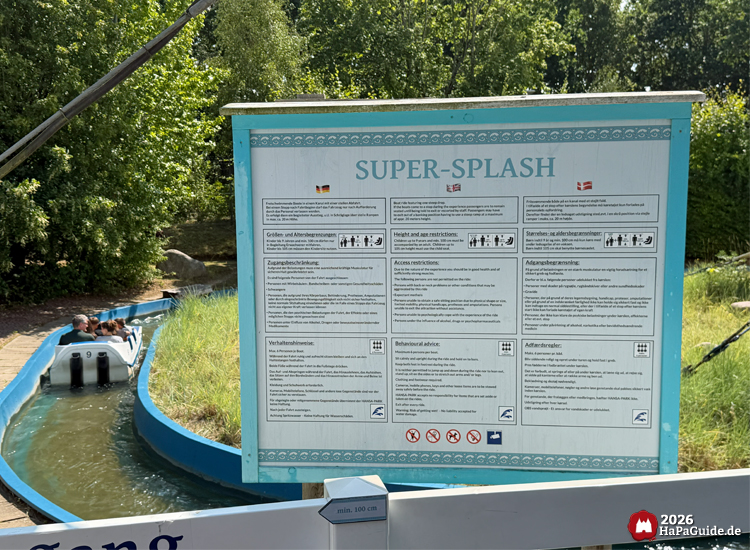 Hinweisschild der Attraktion Super Splash im Hansa-Park mit Sicherheits- und Größeninformationen in Deutsch, Englisch und Dänisch, dahinter Boot im Wasserkanal mit Fahrgästen