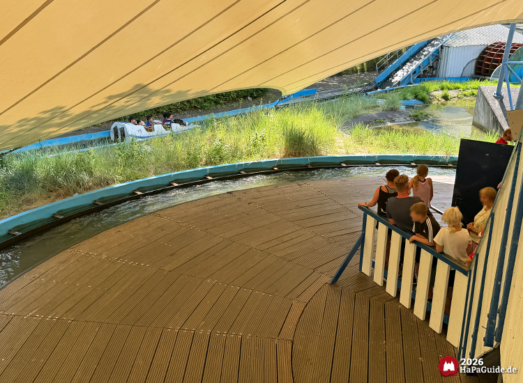 Station der Wasserbahn Super Splash im Hansa-Park unter weißem Zeltdach, Besuchende beobachten Boote im Wasserkanal, umgeben von Gras und drehender Holzplattform