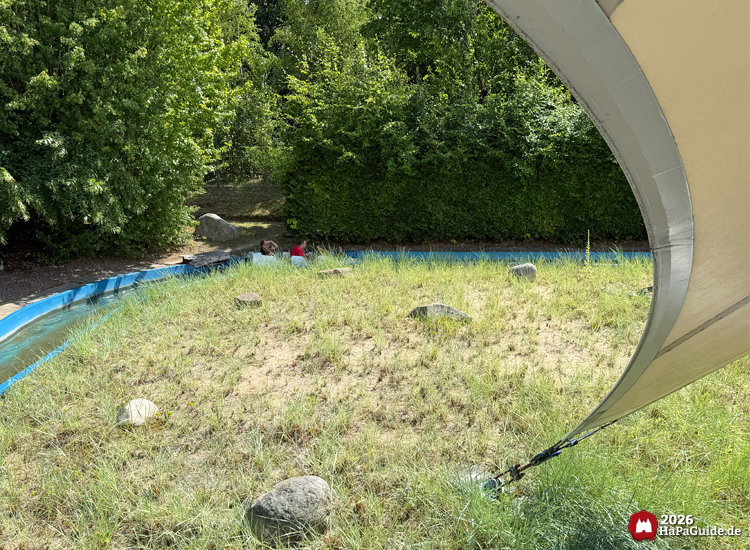 Wasserkanal der Attraktion Super Splash im Hansa-Park mit blauer Rinne und grasbewachsener Insel unter weißem Zeltdach, im Hintergrund dichte grüne Hecken und Bäume