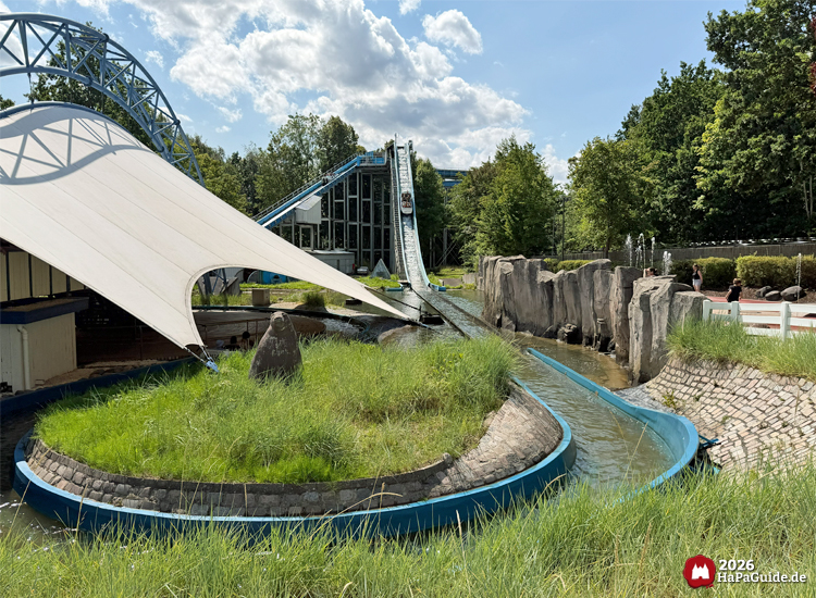 Wasserbahn Super Splash im Hansa-Park mit Boot auf der Wasserabfahrt, blauer Kanal und weißes Zeltdach, umgeben von Felsen und grüner Landschaft unter sonnigem Himmel