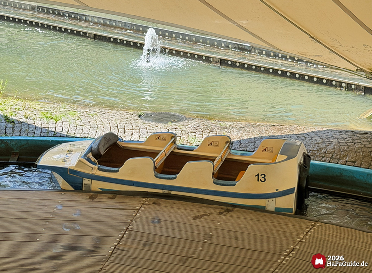 Leeres Boot von Super Splash im Hansa-Park an hölzerner Drehplattform unter weißem Zeltdach, im Hintergrund Wasserkanal mit kleiner Fontäne und Steinmauer