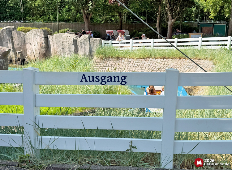 Ausgangsbereich der Wasserbahn Super Splash im Hansa-Park mit weißem Zaun und Schild 'Ausgang', dahinter Wasserkanal mit Boot und Felsenlandschaft im Hintergrund